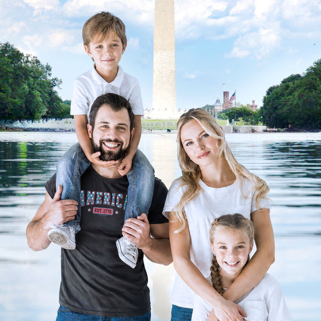 A family of four posing together in front of a Washington Monument backdrop. The father wears a Black t-shirt by America The Beautiful with 'AMERICA EST. 1776' text design
