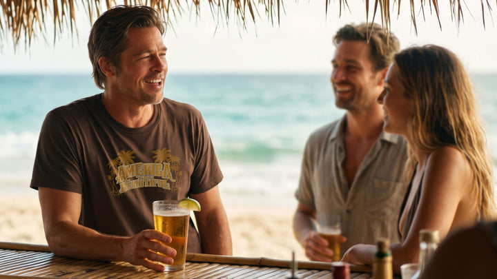 Three people sitting at a beach bar under a thatched roof, laughing and holding drinks. A man wearing a Brown t-shirt with &