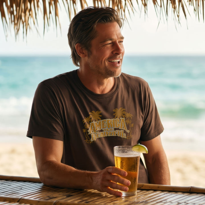 A man sitting at a beach bar under a thatched roof, wearing a Brown t-shirt with &