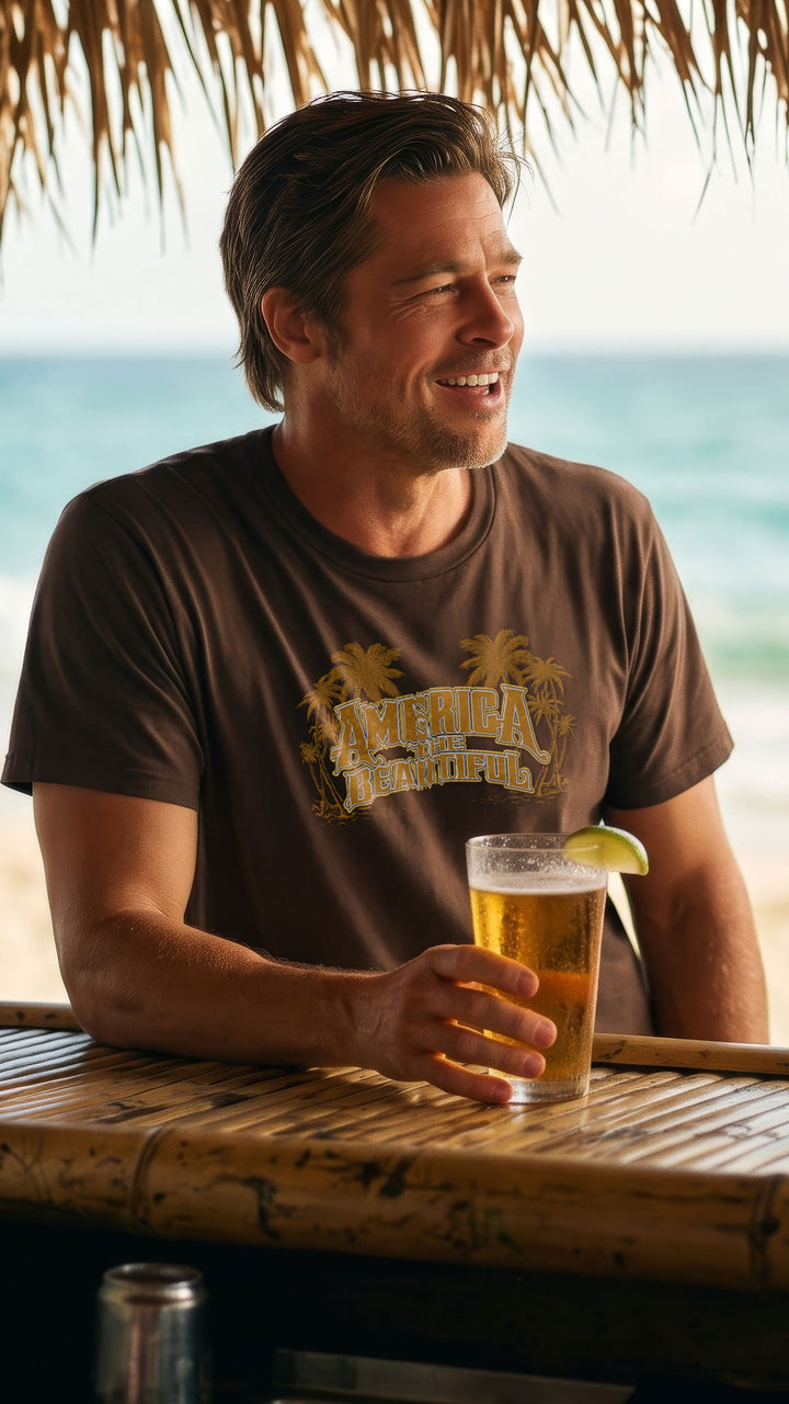 A man sitting at a beach bar under a thatched roof, wearing a Brown t-shirt with &