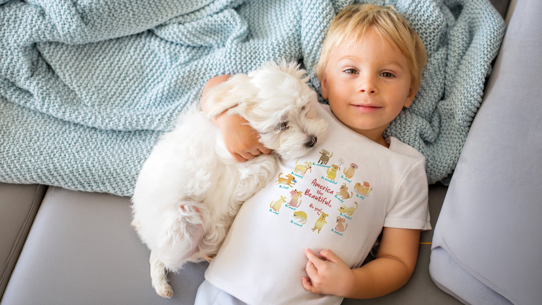 Child holding a small white dog wearing a White t-shirt with colorful "America The Beautiful" with toys graphic design