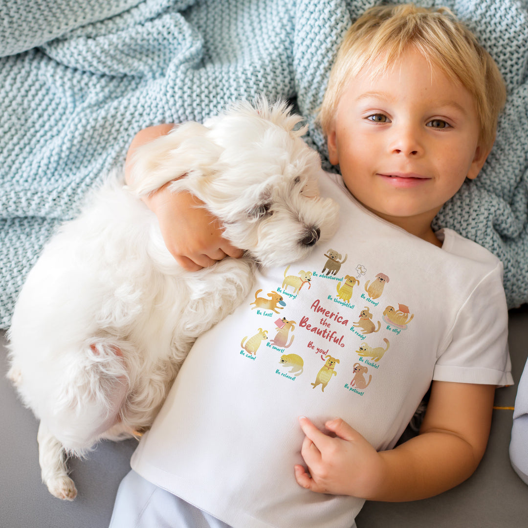 Child holding a small white dog wearing a White t-shirt with colorful "America The Beautiful" with toys graphic design