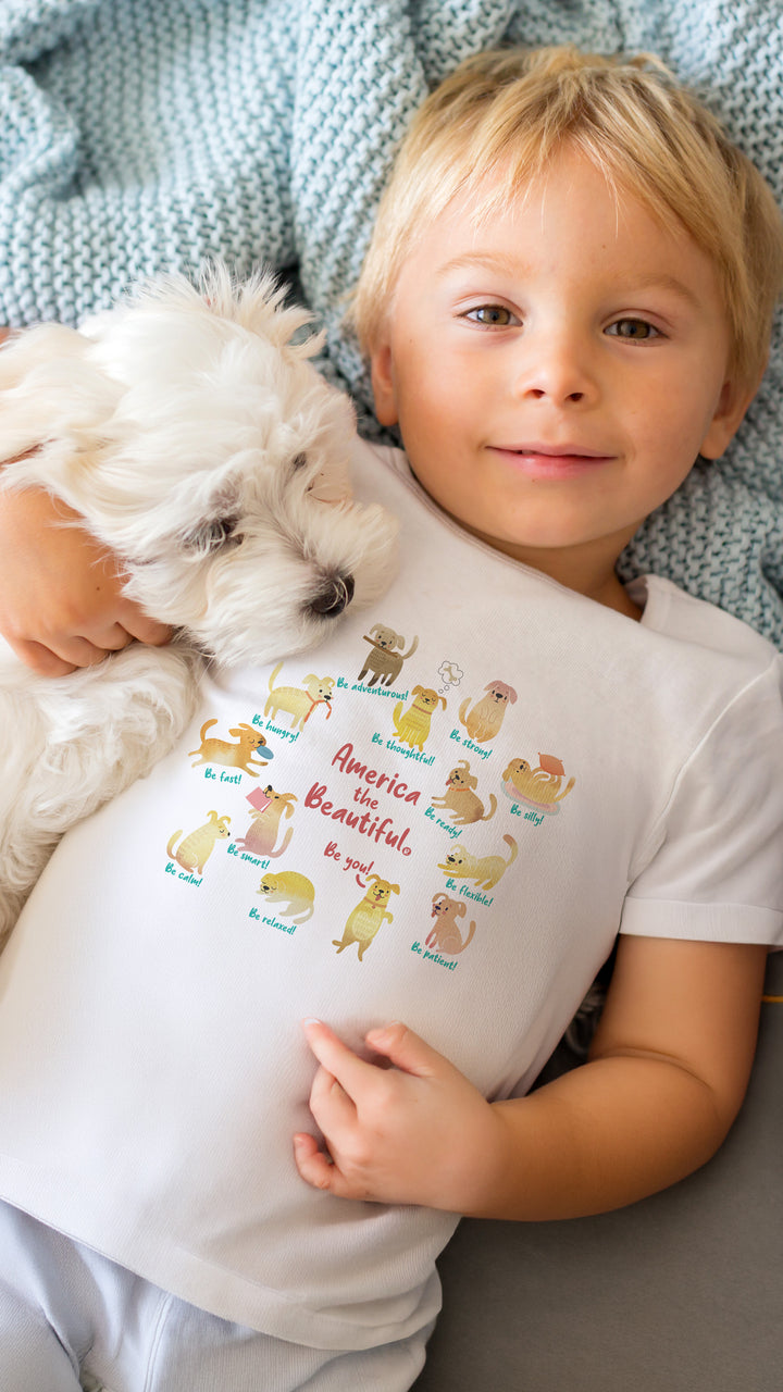 Child holding a small white dog wearing a White t-shirt with colorful "America The Beautiful" with toys graphic design