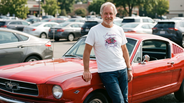 A smiling older man stands outdoors in front of a red 66 Mustang, wearing a white graphic T-shirt with a vintage Mustang headlight design and &