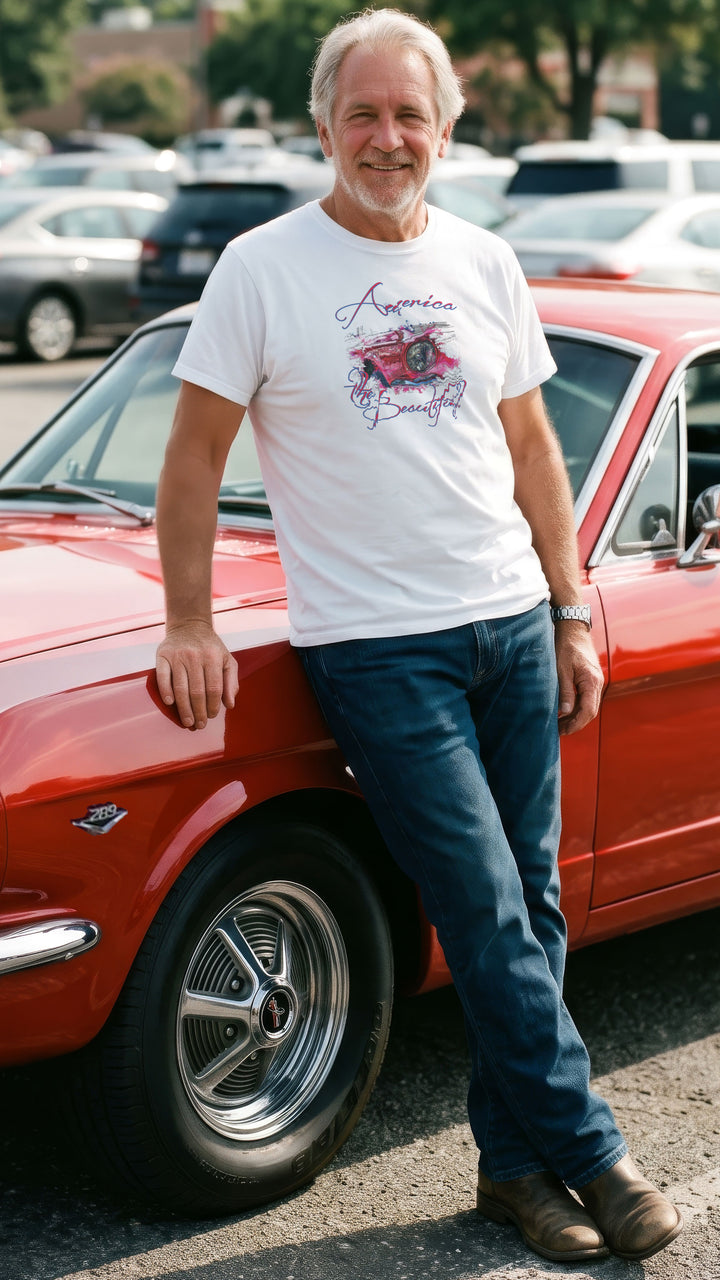 A smiling older man stands outdoors in front of a red 66 Mustang, wearing a white graphic T-shirt with a vintage Mustang headlight design and &