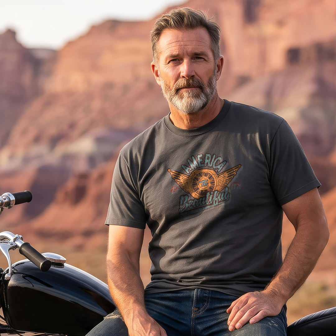 Man leaning on motorcycle and wearing a Gray/green t-shirt with 'America Beautiful' graphic design and a winged motorcycle wheel graphic