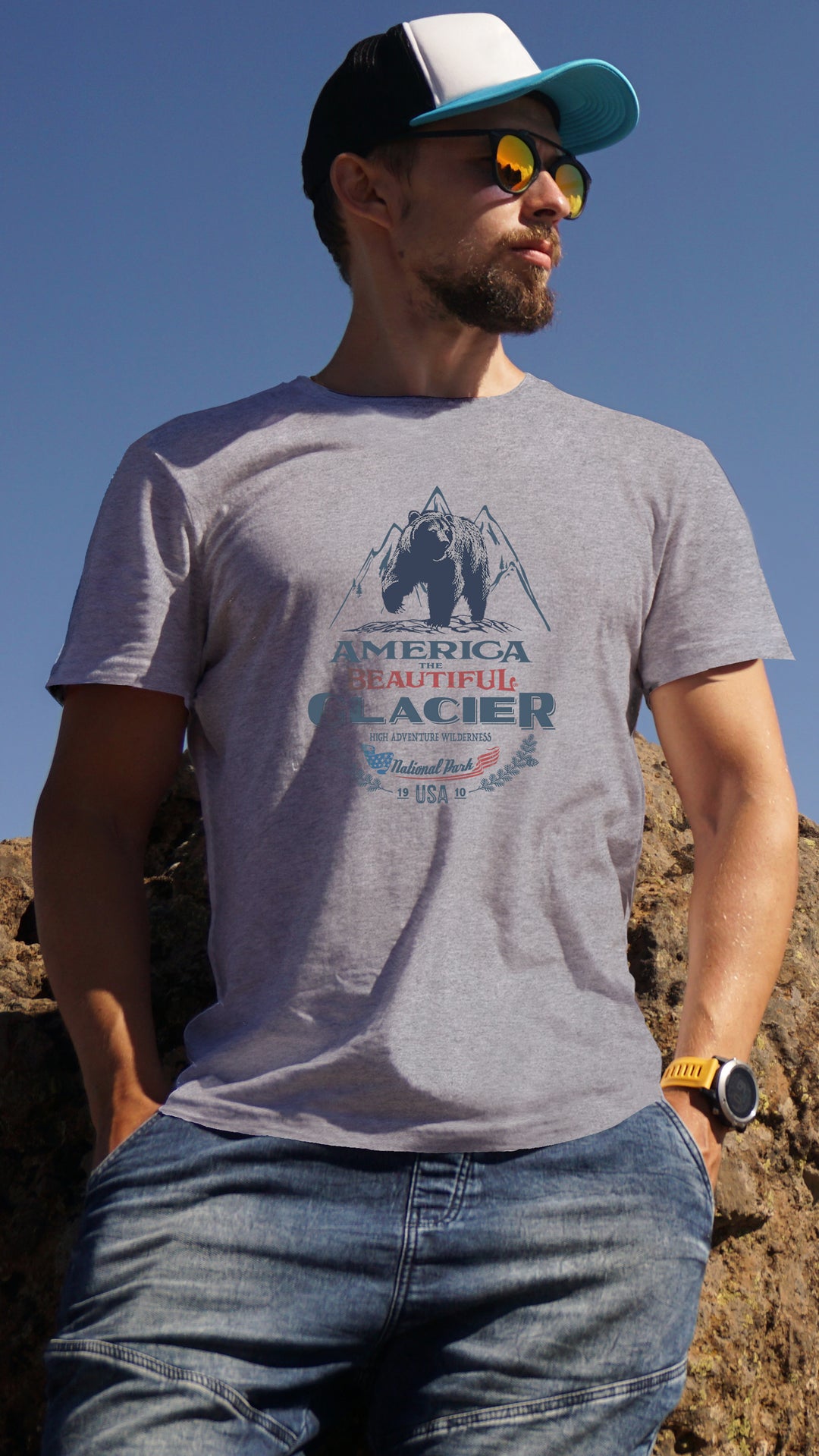 Man wearing a gray America The beautiful t-shirt with a Glacier Park Grizzly Bear outdoors against a clear blue sky.