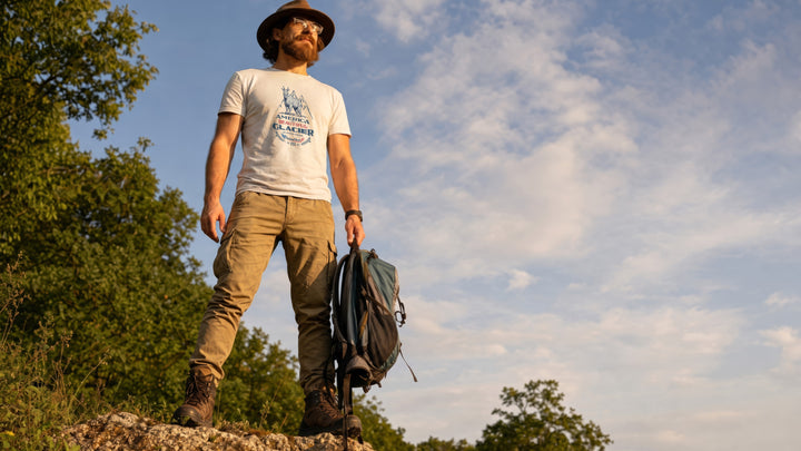 Man wearing a White t-shirt with graphic design featuring a Glacier Nat. Park mountain goat, standing outdoors against a blue sky.