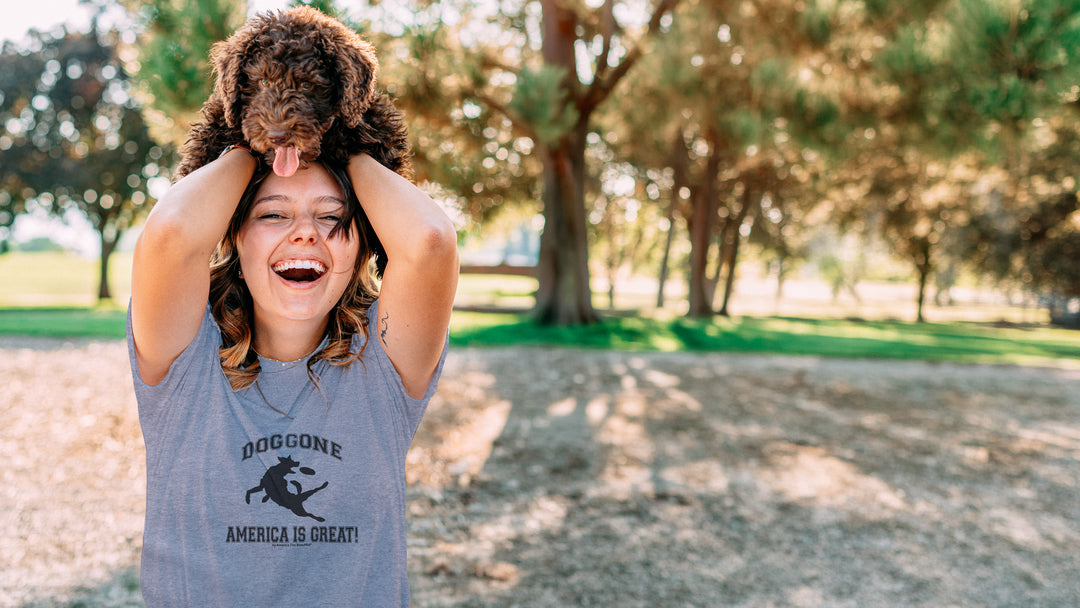 Woman holding a dog on her head in a park, wearing a Gray t-shirt with black dog chasing freesbe graphic and doggone America is great