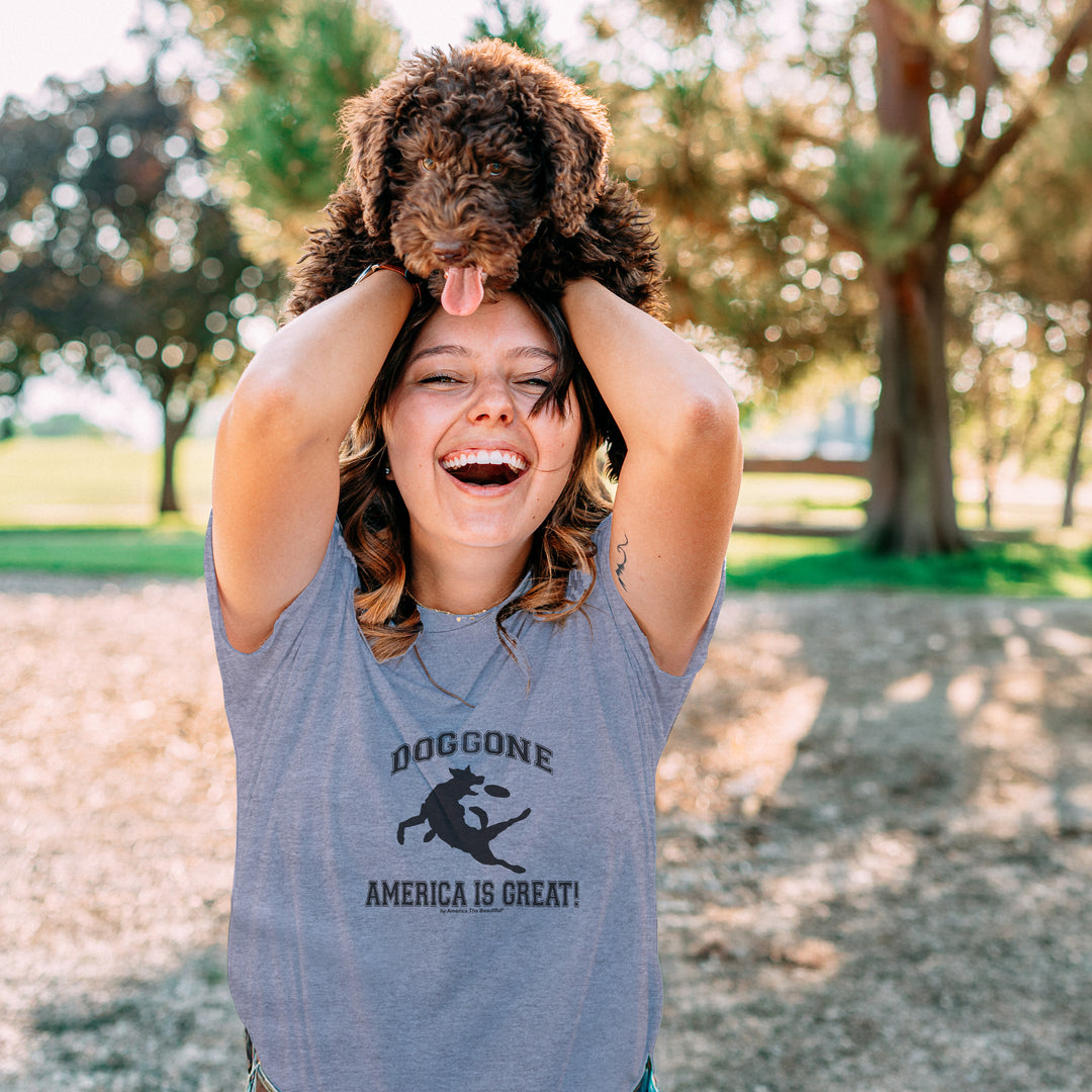 Woman holding a dog on her head in a park, wearing a Gray t-shirt with black dog chasing freesbe graphic and doggone America is great