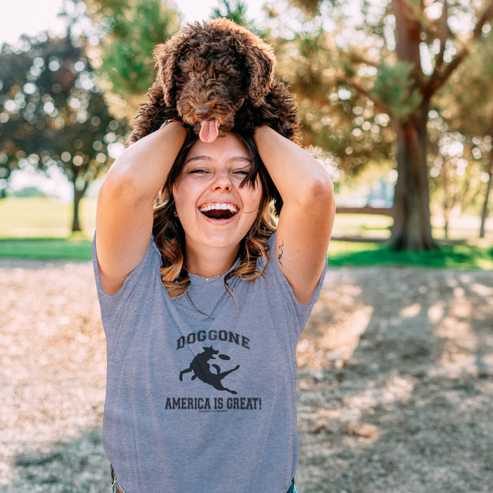 Woman holding a dog on her head in a park, wearing a Gray t-shirt with black dog chasing freesbe graphic and doggone America is great