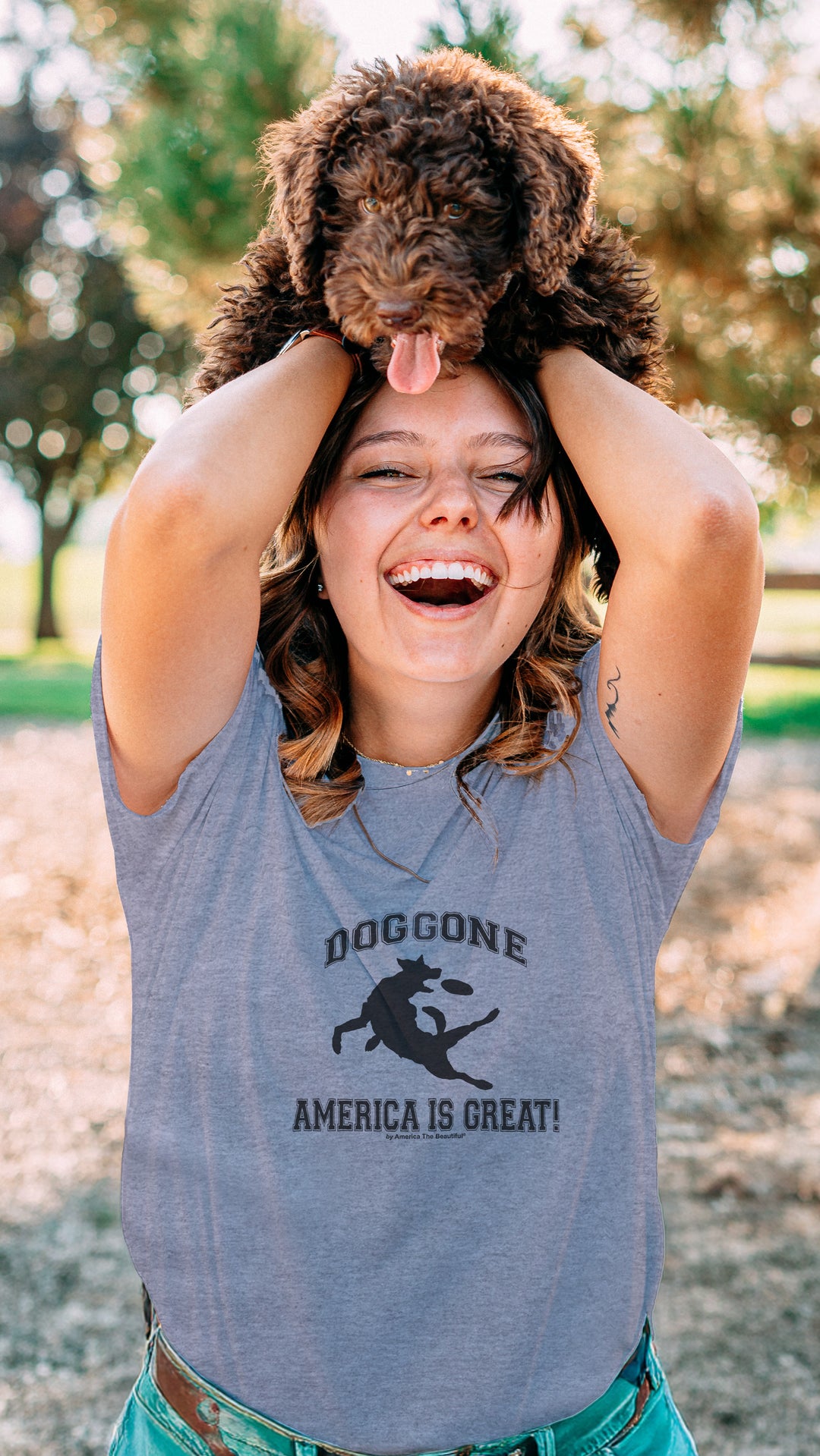 Woman holding a dog on her head in a park, wearing a Gray t-shirt with black dog chasing freesbe graphic and doggone America is great
