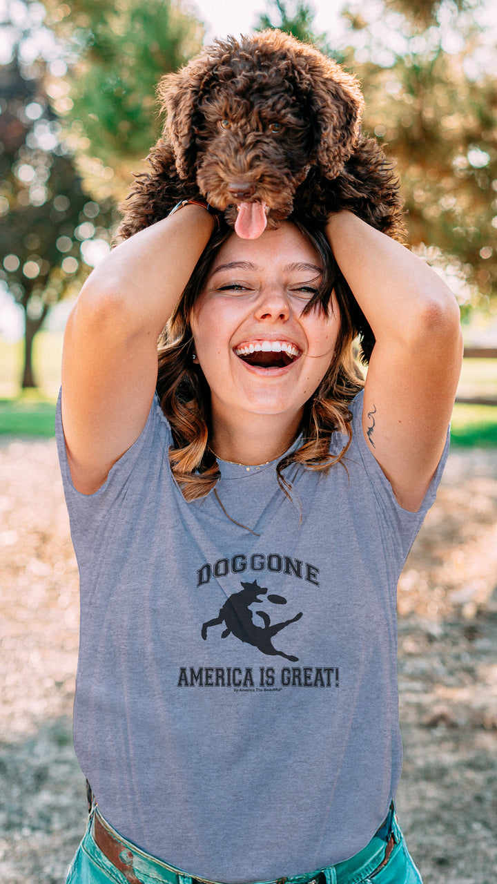 Woman holding a dog on her head in a park, wearing a Gray t-shirt with black dog chasing freesbe graphic and doggone America is great