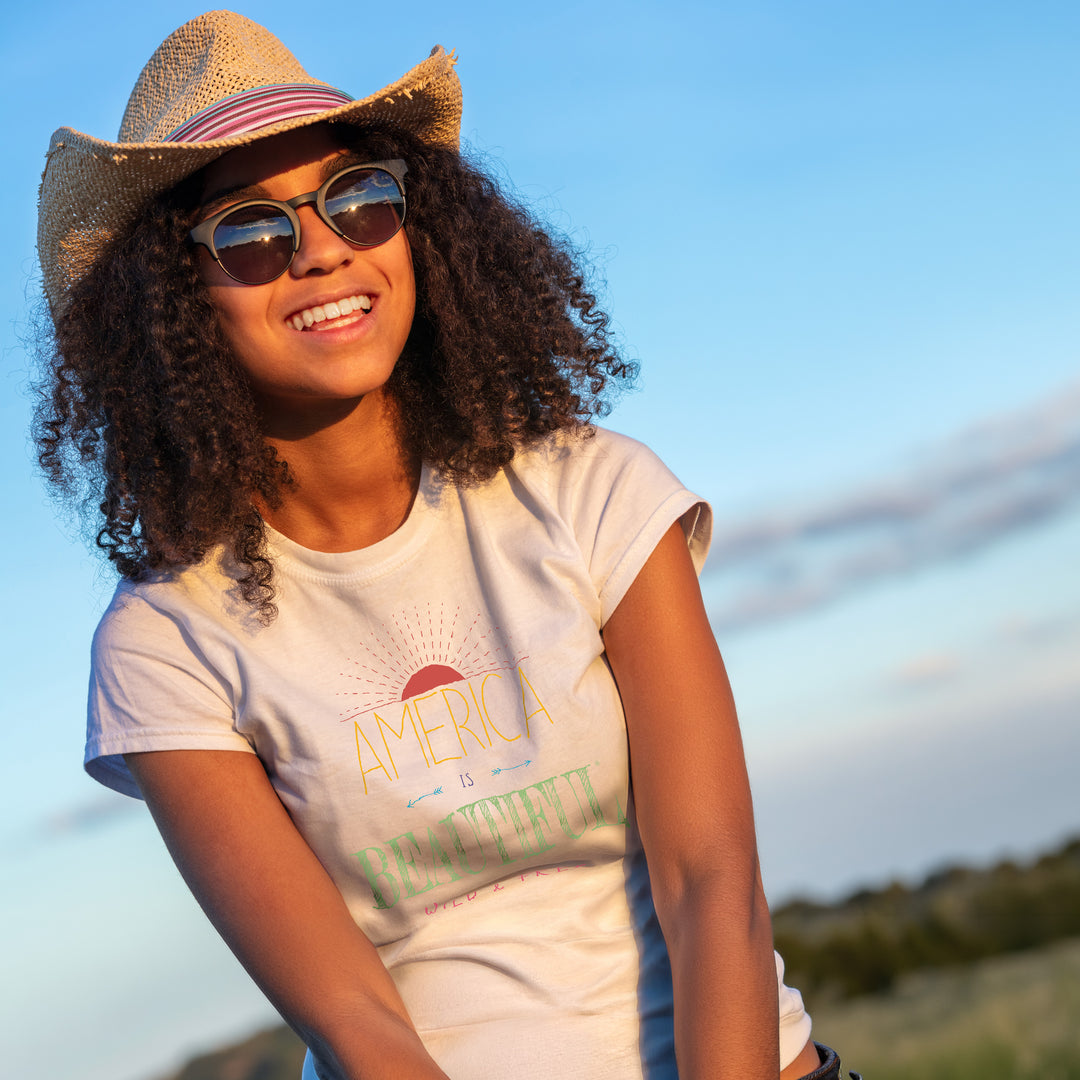 Smiling woman wearing a White t-shirt with 'America is Beautiful, Wild & Free' text under a stylized raising sun