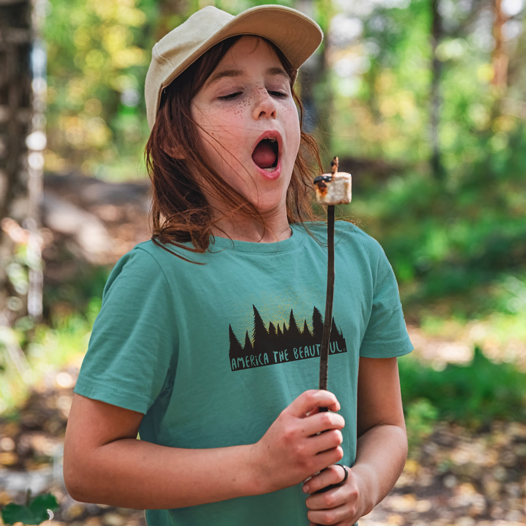 Child roasting a marshmallow wearing a Teal Magical Forest t-shirt with 'America the Beautiful' graphic