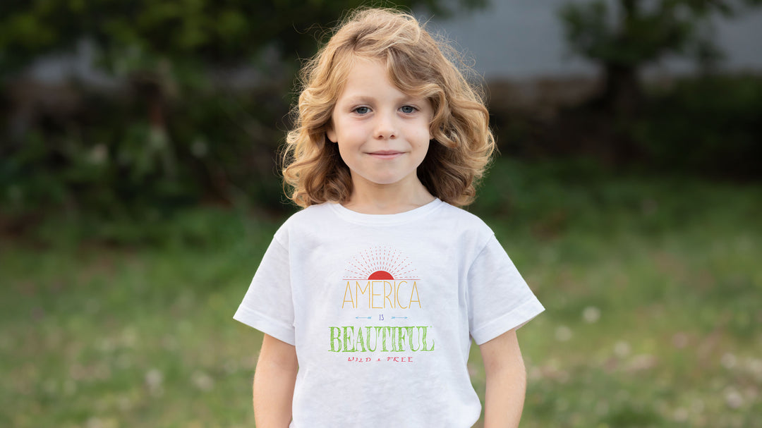 A young girl with curly blonde hair stands outdoors wearing  an America Is Beautiful Wild &amp; Free Sunrise Youth T-shirt design
