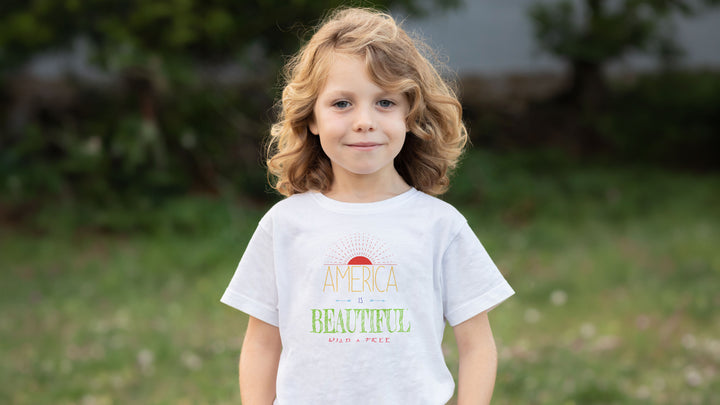 A young girl with curly blonde hair stands outdoors wearing  an America Is Beautiful Wild &amp; Free Sunrise Youth T-shirt design