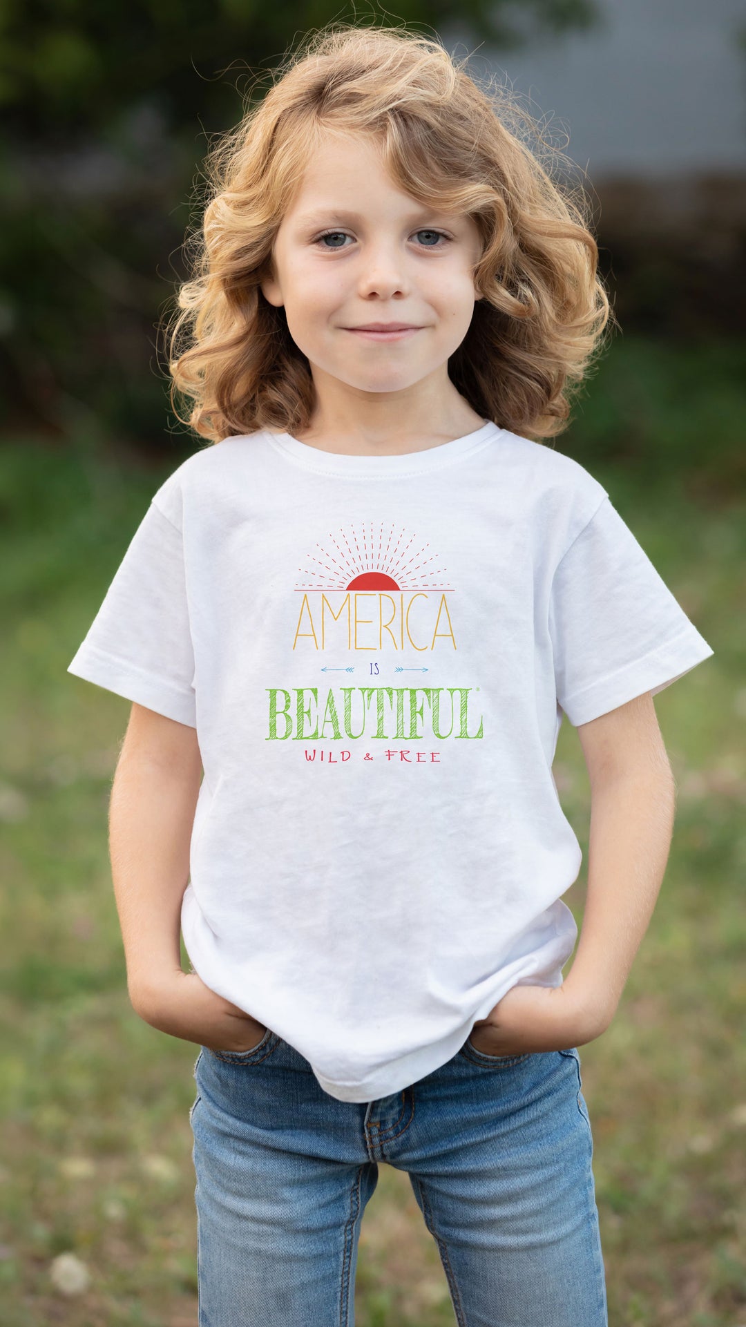 A young girl with curly blonde hair stands outdoors wearing  an America Is Beautiful Wild &amp; Free Sunrise Youth T-shirt design