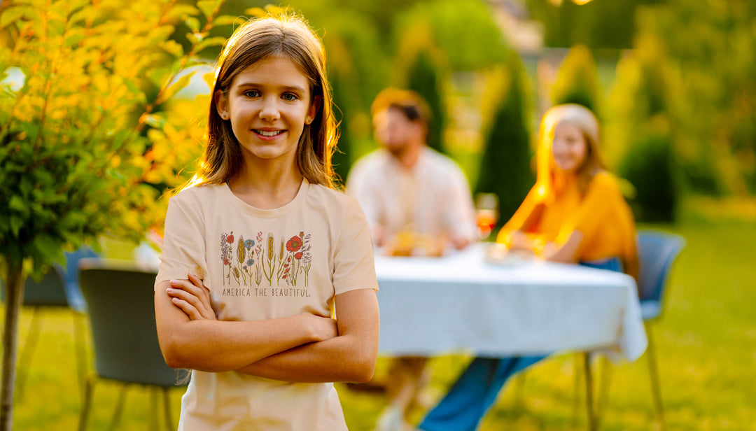 A young girl with long light brown hair stands outdoors with her arms crossed, wearing a light beige graphic t-shirt with a floral design and the text &