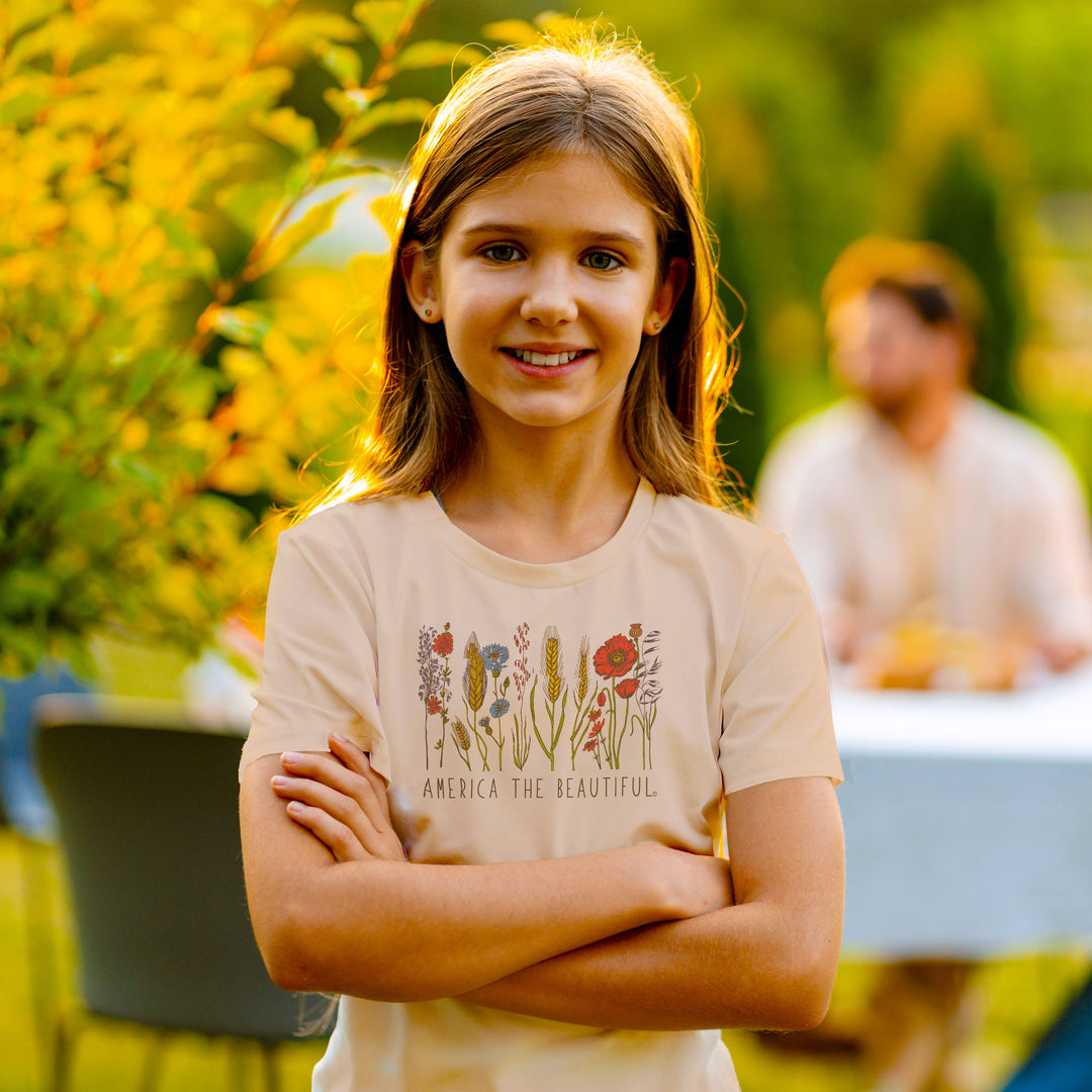 A young girl with long light brown hair stands outdoors with her arms crossed, wearing a light beige graphic t-shirt with a floral design and the text 'AMERICA THE BEAUTIFUL'. The background is a warm, sunlit outdoor setting with blurred greenery and another person in the distance.