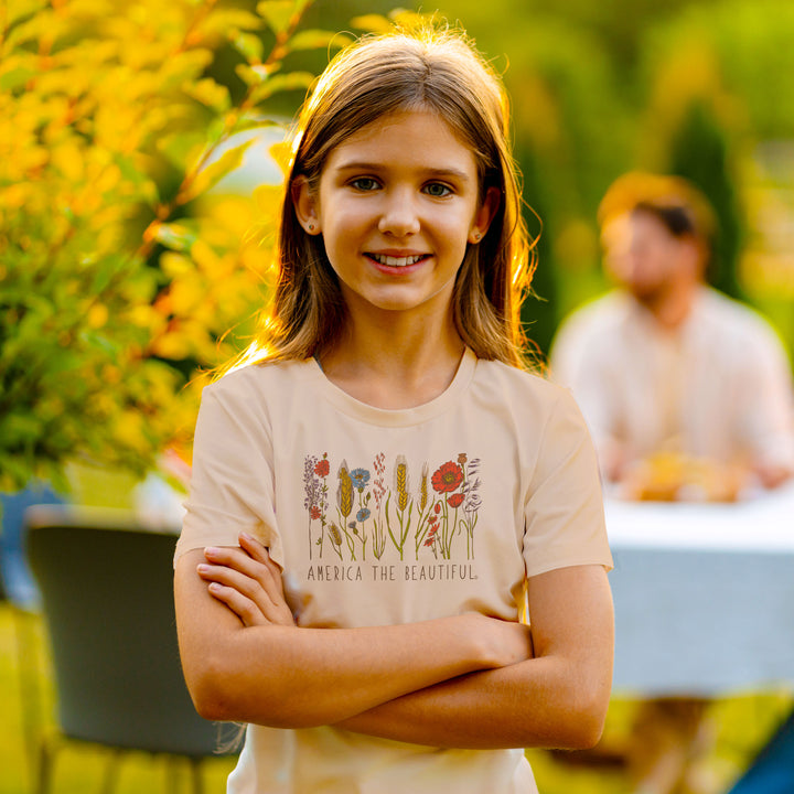 A young girl with long light brown hair stands outdoors with her arms crossed, wearing a light beige graphic t-shirt with a floral design and the text &