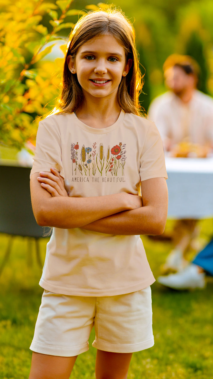 A young girl with long light brown hair stands outdoors with her arms crossed, wearing a light beige graphic t-shirt with a floral design and the text &