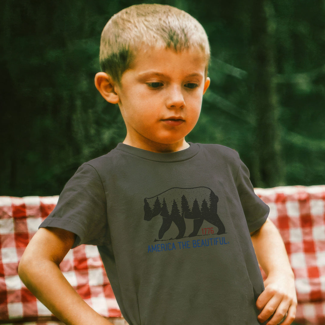 Boy wearing a Military Green t-shirt with bear graphic and 'America the Beautiful' and 1776 text while camping