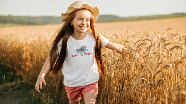 Smiling wild and free Young girl in a field wearing a White t-shirt with wild horses graphic design and America the beautiful text on a white background