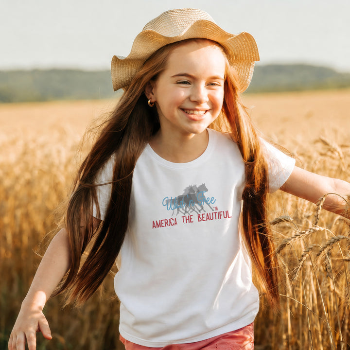 Smiling wild and free Young girl in a field wearing a White t-shirt with wild horses graphic design and America the beautiful text on a white background