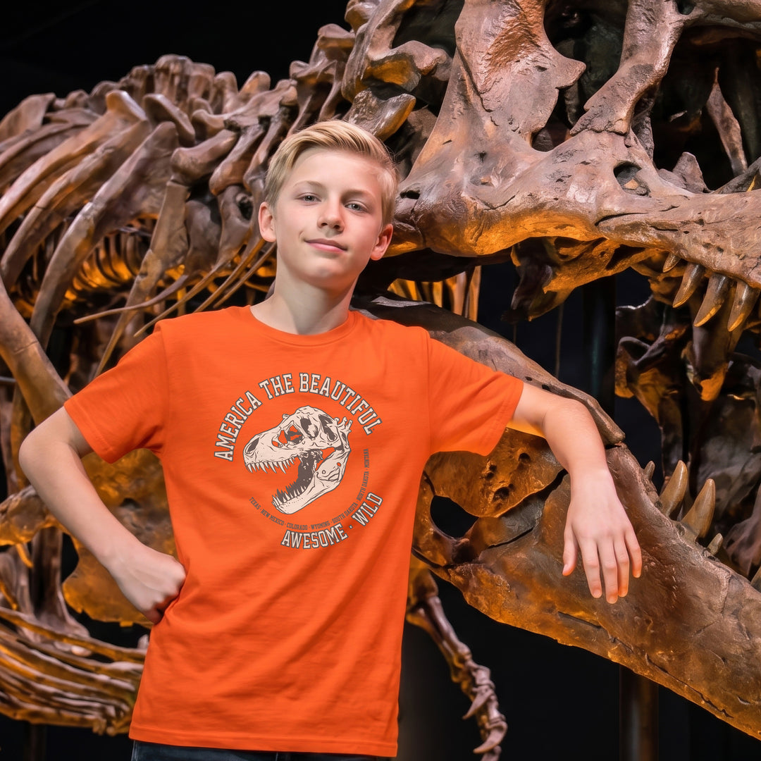 Boy wearing an orange t-shirt with a T-rex dinosaur graphic in front of a dinosaur skeleton display