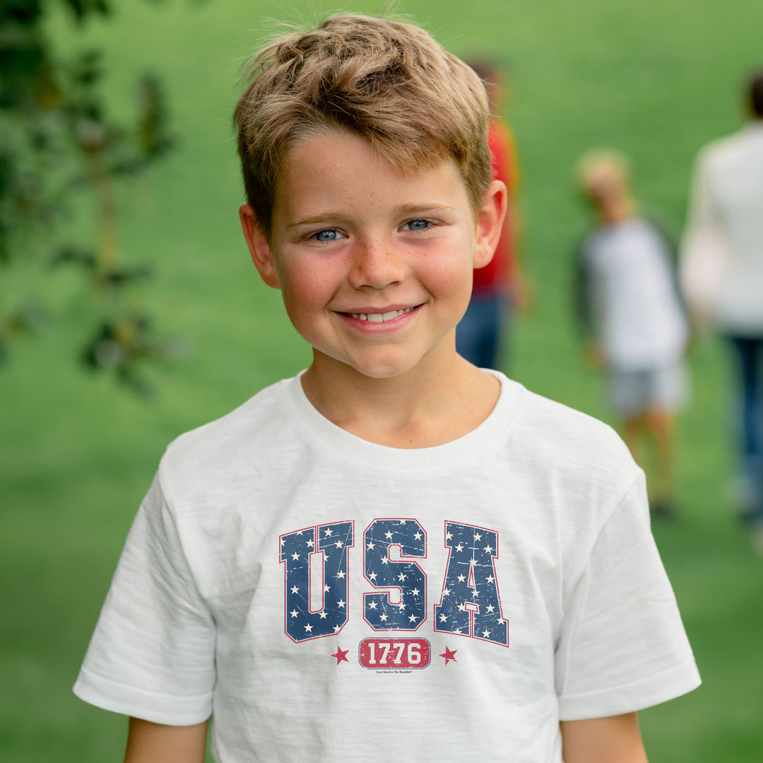Young boy wearing a white t-shirt with 'USA 1776' printed on it, standing outdoors with green grass in the background.