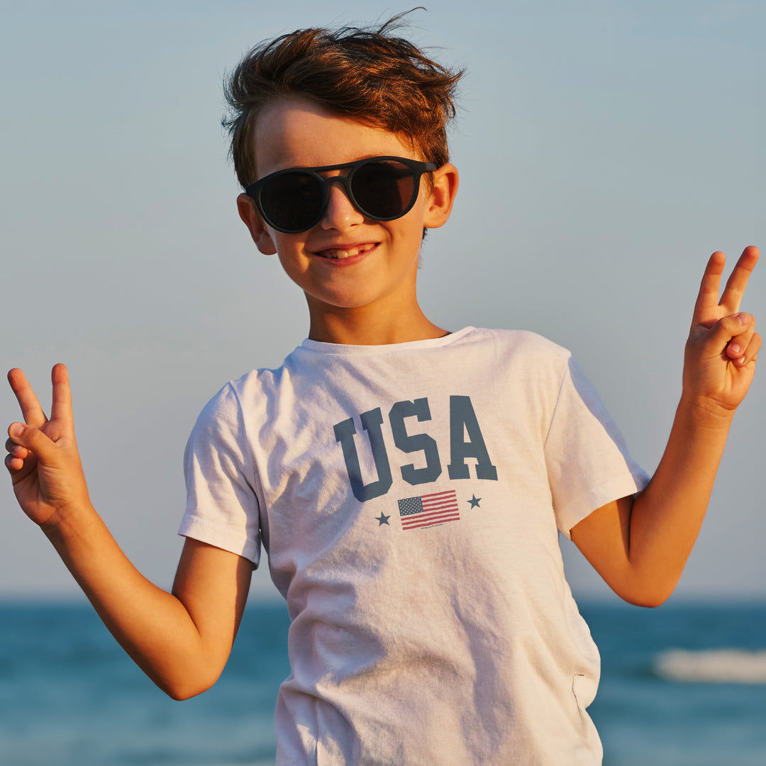 Child wearing sunglasses and a 'USA' t-shirt with an "America the beautiful" American flag against a clear sky.