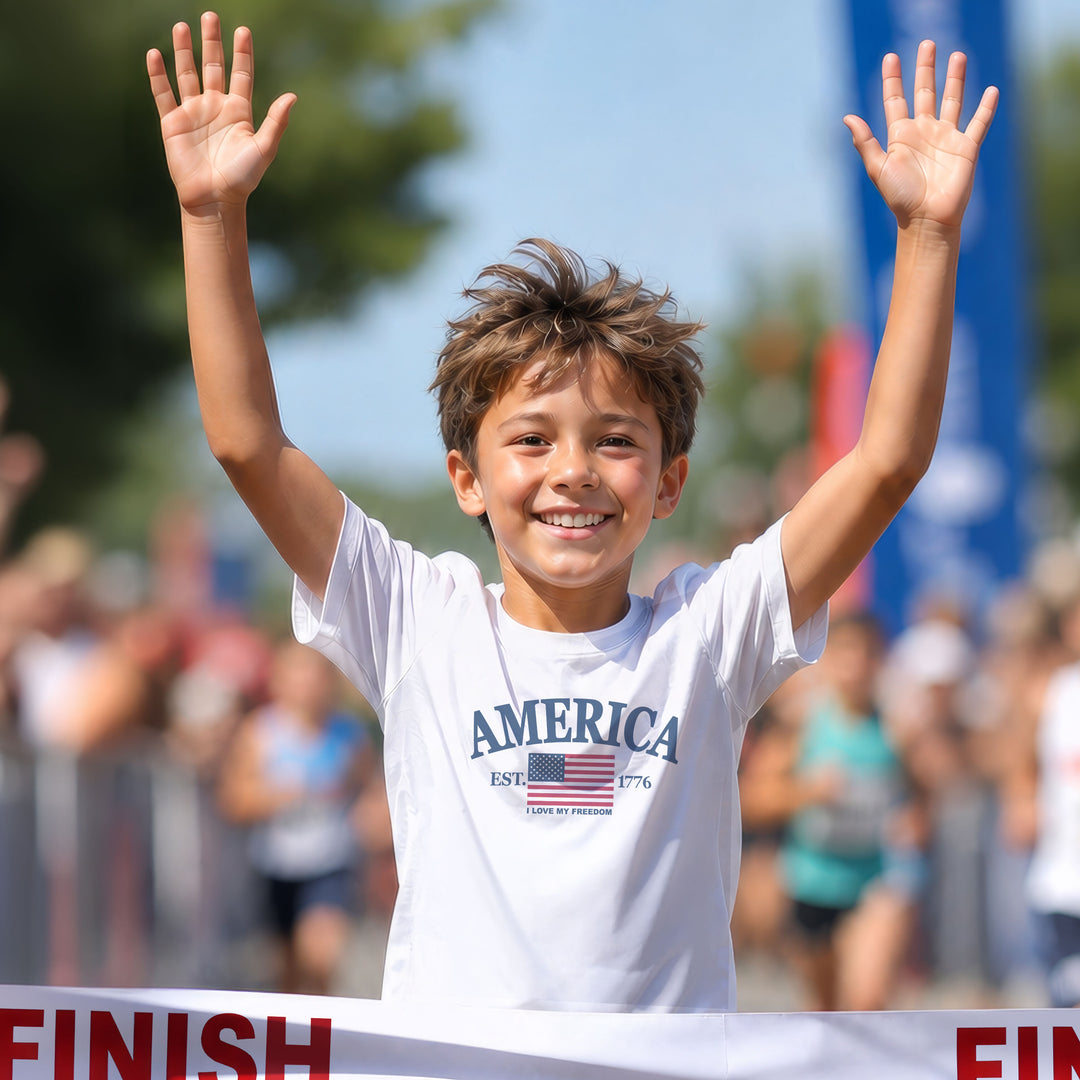 Running young boy wearing a white t-shirt with 'AMERICA, est. 1776' and 'Love my freedom' with an American flag design, crossing a race finish line.