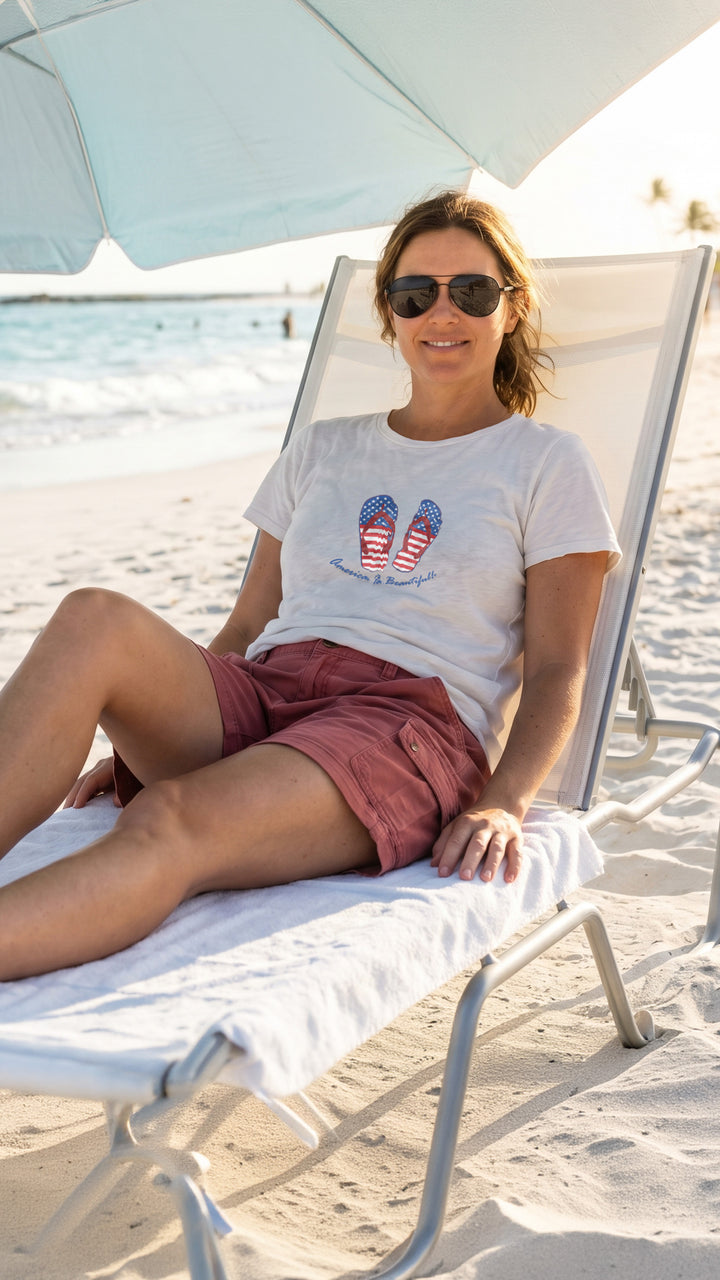 Woman sitting on a beach chair under an umbrella, wearing sunglasses and a t-shirt with a red white and blue graphic design of flip flops.